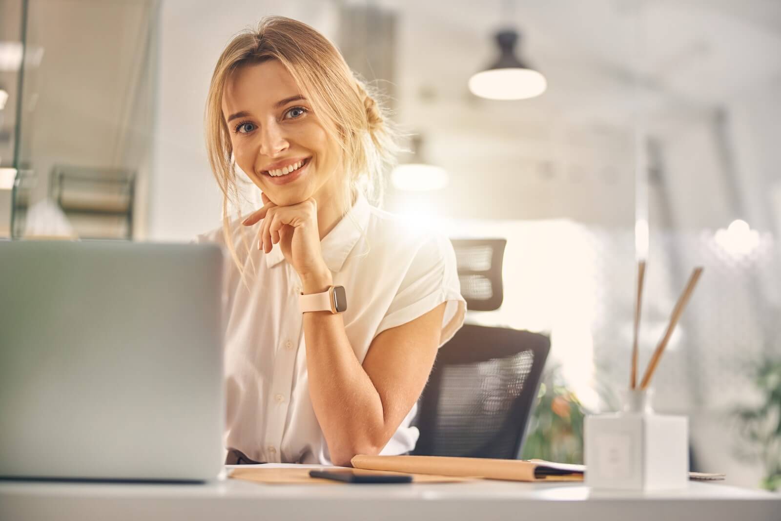 charmante-blonde-dame-die-laechelt-waehrend-sie-mit-laptop-bei-der-arbeit-am-tisch-sitzt